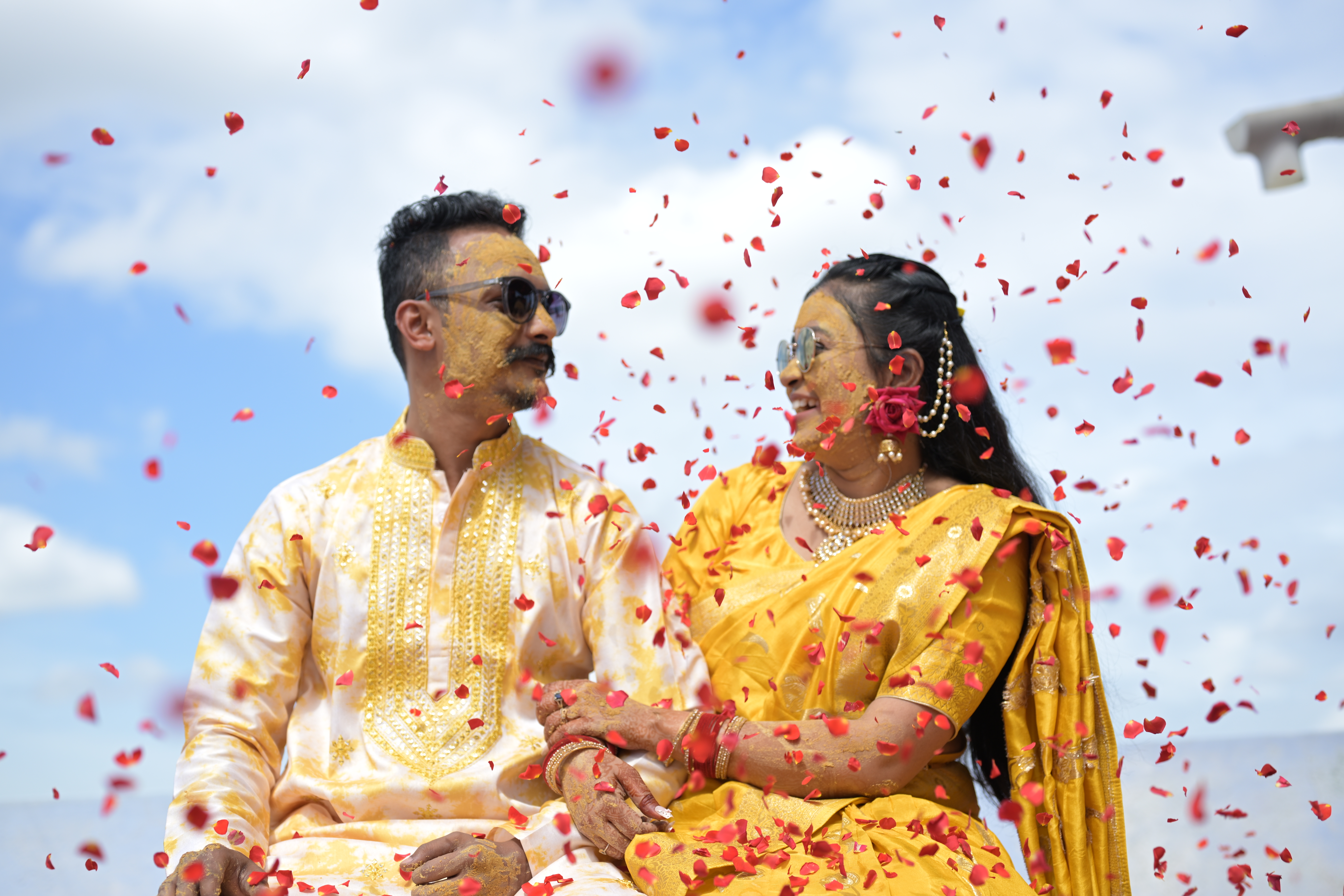 Couple in traditional yellow wedding attire laughing together under falling rose petals